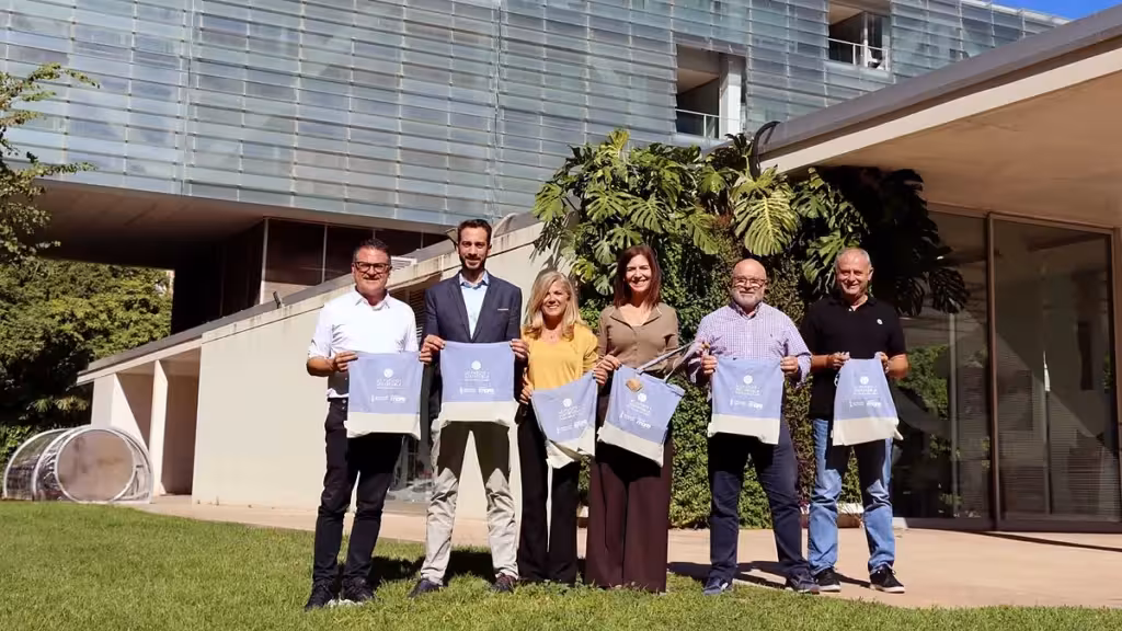 Seis personas sostienen bolsas de la campaña de reciclaje delante de un edificio con vegetación.