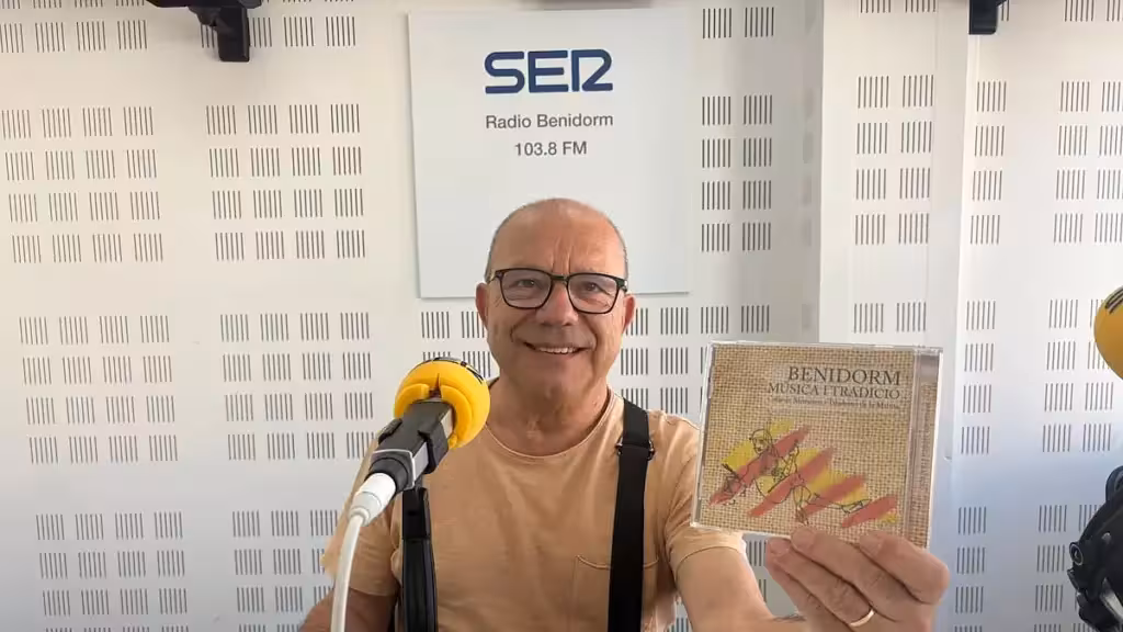A man holds up the Benidorm Música i Tradició CD beside a microphone in a radio studio