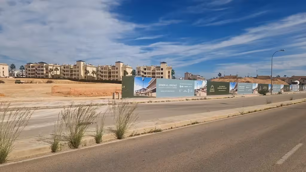 Solar en obras y vallado publicitario de Campo Maximo junto a una calle y edificios residenciales de Playa Flamenca