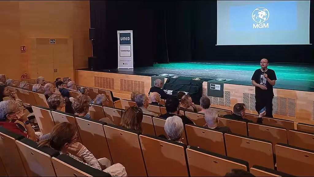 Público en un auditorio ante un ponente con micrófono y una pantalla de cine, junto a un expositor de UNED Torrevieja