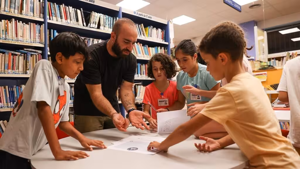 Niños y un adulto trabajan con hojas sobre una mesa entre las estanterías de una biblioteca.