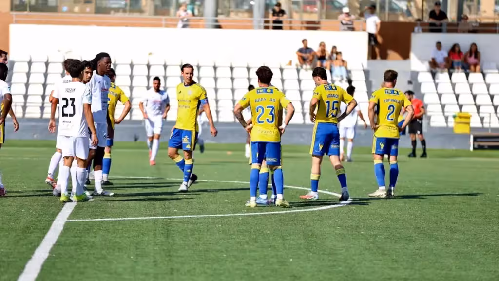 Jugadores con equipaciones amarillas y azules y blancas sobre el césped, ante una grada de asientos blancos