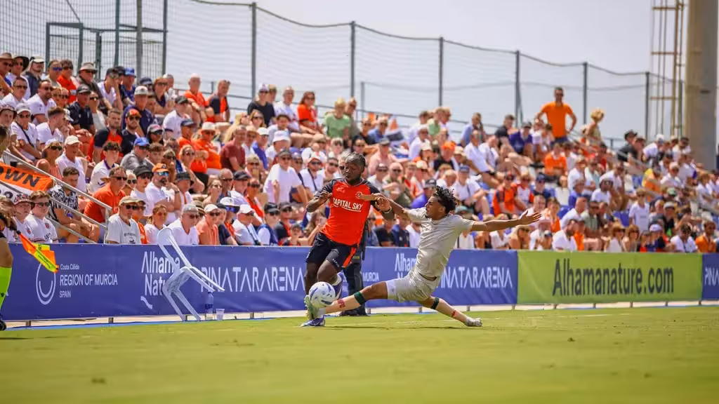 Dos futbolistas disputan el balón junto a una grada llena y vallas publicitarias de Pinatar Arena