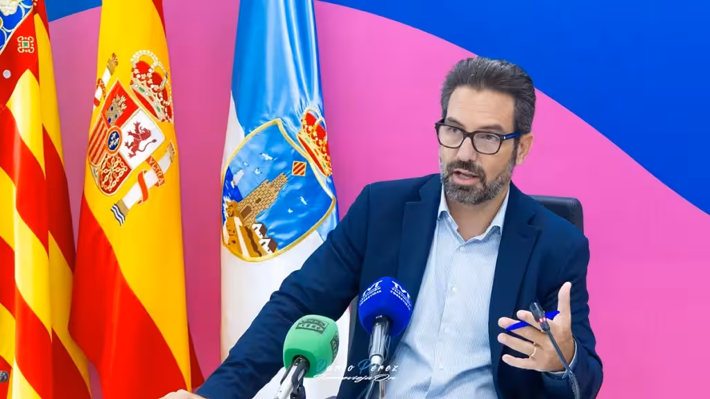 A man speaking into microphones beside institutional flags